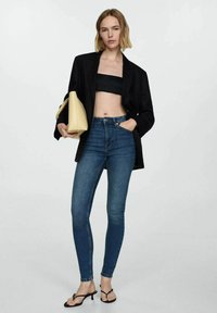 Črn oversized blazer, črn kratki top, temno modre skinny jeans hlače, rumena torbica, črne sandale s peto, gladke teksture, prilegajoča oblika.