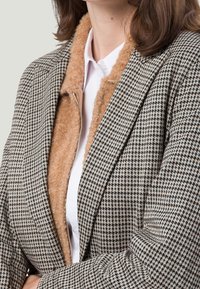 Teksturowana marynarka w zygzaki w kolorach beżowym i czarnym z miękkim, jasnobeżowym kołnierzem z futra. Posiada zamek błyskawiczny i dopasowany krój. Pod spodem biała koszula.