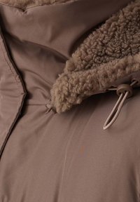 Brązowa odzież wierzchnia z miękkim, teksturowanym beżowym kołnierzem, wyposażona w troczki oraz gładką, matową tkaninę. Projekt jest smukły i nowoczesny.