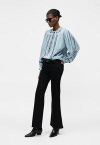 Γυναίκα με υπερμεγέθη γαλάζιο κεντητό πουκάμισο, μαύρο καμπάνα παντελόνι, μαύρη ζώνη, μαύρα μποτάκια και μαύρα γυαλιά ηλίου, όρθια στο πλάι.