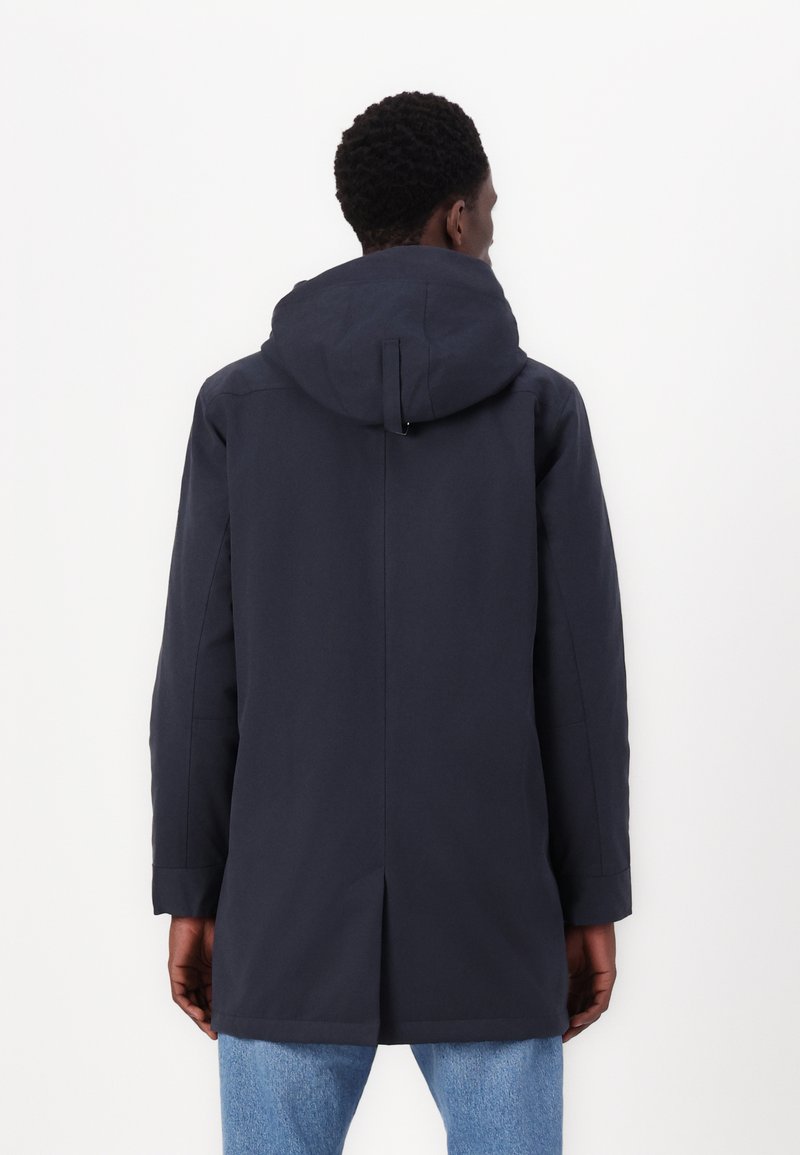 Ciemnoniebieska kurtka z kapturem o gładkim, matowym wykończeniu, z centralnym wentylem i długimi rękawami, noszona na jasnoniebieskich dżinsach.