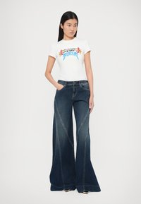 Biele grafické tričko s farebnou potlačou, skombinované s tmavomodrými džínsami s širokými nohavicami, ktoré majú jedinečné švové detaily a bočné vrecká.