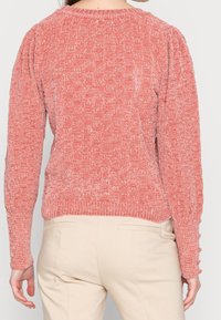 Pull en maille rose texturée avec col rond, manches bouffantes et ourlet côtelé. Présente des boutons décoratifs le long des poignets des manches.