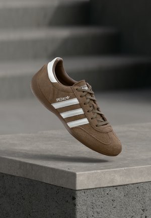 Brązowy zamszowy sneaker Adidas Spezial z białymi paskami i sznurowadłami, unoszący się nad szarą betonową krawędzią na tle rozmytych schodów.