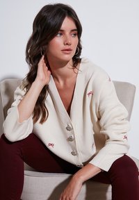 Cardigan color crema realizzato in maglia testurizzata, con quattro bottoni argentati e dettagli in ricamo rosso sulle maniche.