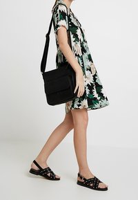 Kvinna som går i en blommig klänning, bär svarta nitar sandaler och har en svart axelväska över axeln.