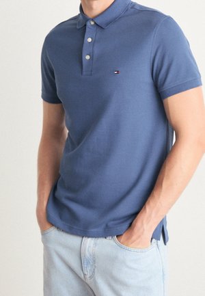 Man draagt een blauw polohemd met korte mouwen en lichtblauwe spijkerbroek met handen in de zakken tegen een effen achtergrond.