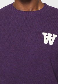 Pull tricoté violet avec une texture côtelée, présentant un logo blanc "W" sur le côté gauche de la poitrine. Design classique à col rond.