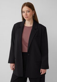 Zwarte blazer met een enkele knoop, notch revers en zijzakken, gelaagd over een mauve t-shirt. Glad materiaal met een ontspannen pasvorm.