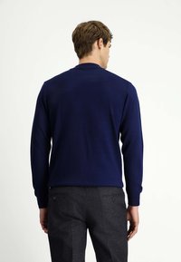 Granatowy sweter dzianinowy z długimi rękawami, okrągłym dekoltem i prążkowanymi mankietami, noszony na szarych spodni z wzorem.