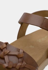 Καφέ δερμάτινο σανδάλι με υφαντό σχέδιο στο λουράκι των δακτύλων, λεία επιφάνεια και επίπεδη σόλα με εσωτερική σόλα σε ανοιχτό καφέ χρώμα.