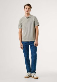 Polo shirt gris clair avec un col classique, manches courtes, porté avec un jean bleu et des baskets blanches avec des accents noirs et une semelle en caoutchouc.