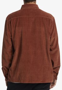 Brown corduroy shirt featuring vertical ribbing, a collar, and buttoned cuffs. The back showcases a relaxed fit with subtle stitching details.