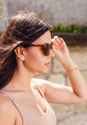 Sunglasses with a tortoiseshell pattern, dark lenses, and a thin frame. Model wearing a light pink top, holding the frame near her face.