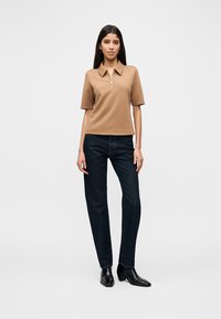 Καφέ κοντομάνικη πόλο μπλούζα με γιακά και μισή φερμουάρ, συνδυασμένη με σκούρο μπλε τζιν παντελόνι ίσιας γραμμής και μαύρα μποτάκια έως τον αστράγαλο.