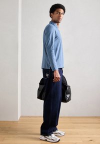 Helehall sinine pikkade varrukatega polo särk, tumelilla teksapüksid taskul oleva logoga ja must tekstuuriga duffel-kott, kantud valgete tossudega.