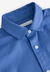 Camicia azzurra con colletto classico, bottoni bianchi e tessuto testurizzato. L'etichetta riporta "NEXT", prodotta in India.