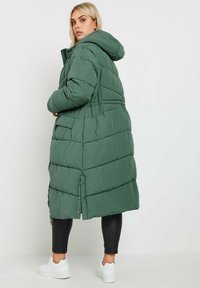 Grön puffjacka med huva, med en kviltad design, tryckknappar och midjeintryck, kombinerad med svarta leggings och vita skor.