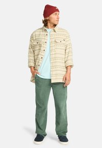 Chemise beige à manches longues avec des motifs vert olive, t-shirt bleu clair, pantalon en velours côtelé vert et bonnet bordeaux avec un insigne. Chaussures marines.
