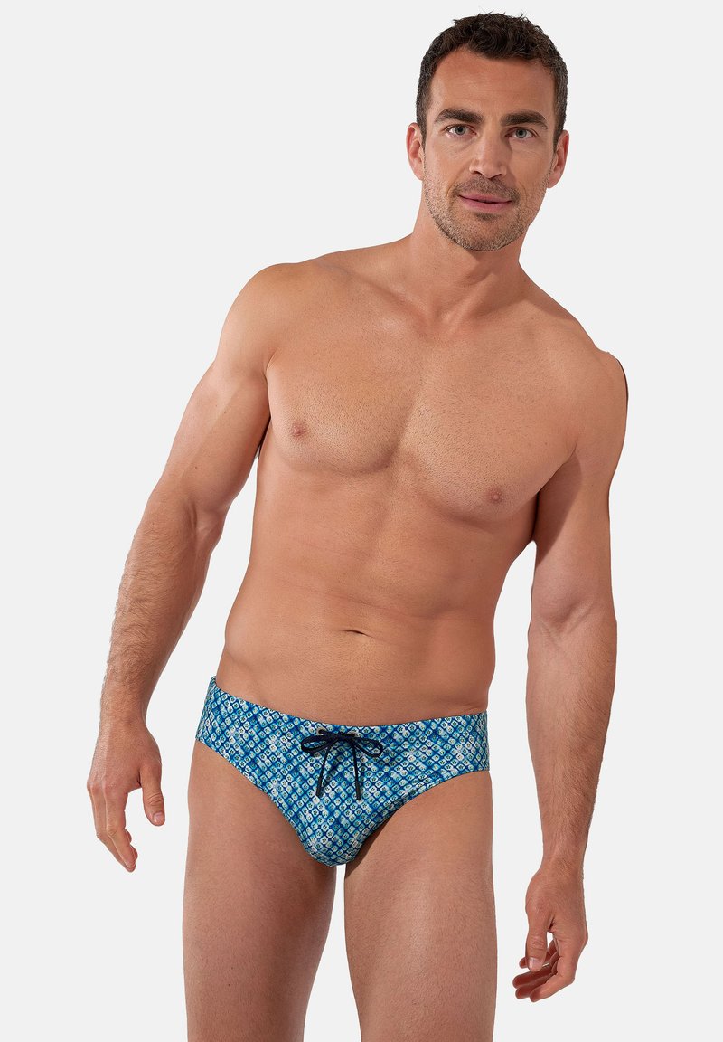 Homme sportif portant un short de bain bleu à motifs avec un cordon noir, debout devant un fond uni clair.