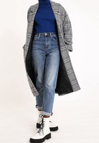 Cappotto lungo a quadri sopra un dolcevita blu, abbinato a jeans azzurri e stivali bianchi con lacci e suole nere.