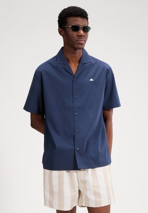 Muškarac u tamno plavoj košulji s kratkim rukavima s gumbima i bež-bijelim kratkim hlačama s uspravnim prugama, s naočalama za sunce, pozira ispred jednobojne pozadine.