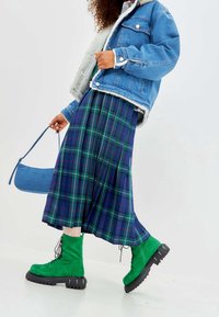 Chaqueta de mezclilla con forro de felpa, falda midi a cuadros en azul y verde, y botas de ante verdes con cordones y suelas gruesas. Acompañada de un bolso azul.