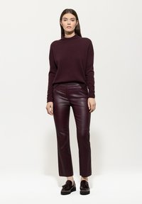 Eine Frau steht in einem dunkelvioletten Rollkragenpullover, passenden Lederhosen und dunklen Quastenloafern vor einem weißen Hintergrund.