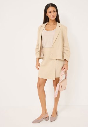 Beige linen blazer over a matching fitted skirt. Light tank top, blush handbag with floral accent, and taupe slingback heels. Smooth texture.