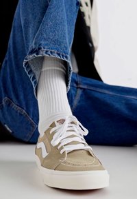 Sneaker beige con lacci bianchi e logo laterale, caratterizzati da una texture liscia e una suola resistente. Indossati con calzini leggeri a coste e jeans blu.