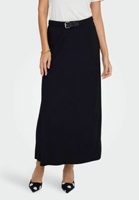 Jupe noire longueur genou en matériau doux, dotée d'une taille ajustée avec une ceinture. Associée à des escarpins à pois noirs et blancs.