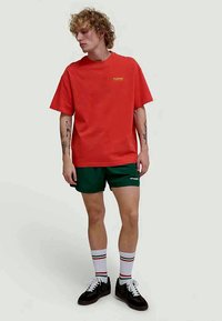 Rode katoenen t-shirt met een gele logo, gecombineerd met groene shorts. Model draagt witte sokken met gekleurde strepen en zwarte schoenen.