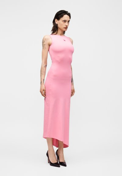 Γυναίκα με τατουάζ που φοράει ροζ εφαρμοστό midi φόρεμα χωρίς μανίκια και μαύρες ψηλές γόβες, στέκεται μπροστά σε ένα απλό φωτεινό φόντο.