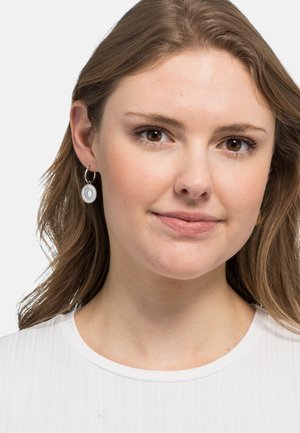 Silberne Creolen mit einem runden, hängenden Anhänger, der konzentrische Ringe zeigt. Das Modell hat helles Haar und trägt ein geripptes weißes Top.