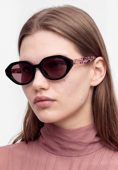 Femme aux cheveux lisses châtain clair porte de grandes lunettes de soleil à monture noire avec des branches roses à motif léopard et un haut mauve à col montant.