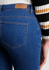 Jeans de mezclilla en azul oscuro, con una cintura alta, costuras en los bolsillos traseros y una etiqueta de cuero marrón con el nombre de la marca "ONLY."