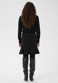 Black sweater and flared black skirt worn with knee-high black pointed boots. Smooth fabric with a subtle texture. Back view.