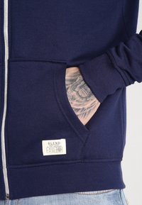Marine blauwe rits hoodie van zachte stof, met zijzakken en een label aan de zoom. De manchetten hebben een ribbeltextuur.