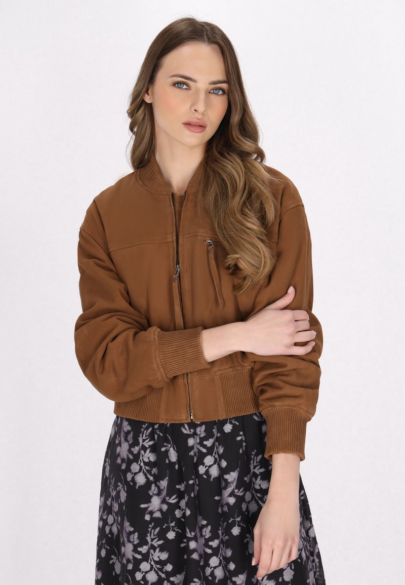 Vrouw met lang golvend haar, gekleed in een bruine zip-up jas en een zwarte rok met een grijs bloemmotief, met de armen over elkaar op een witte achtergrond.