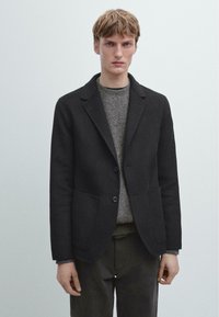 Czarna wełniana marynarka z kołnierzem z wycięciem, zapinana na jeden guzik, z dwoma przednimi kieszeniami, noszona na teksturowanym szarym swetrze.