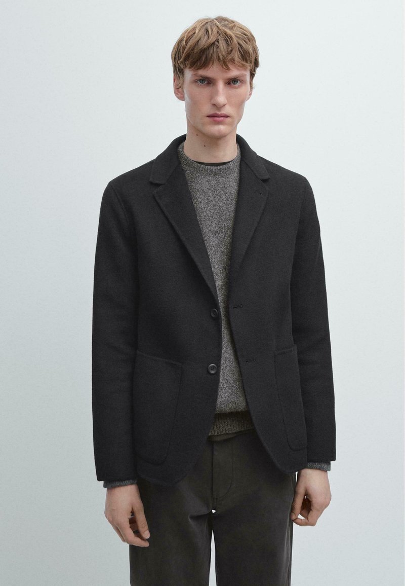 Czarna wełniana marynarka z kołnierzem z wycięciem, zapinana na jeden guzik, z dwoma przednimi kieszeniami, noszona na teksturowanym szarym swetrze.