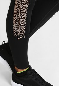 Leggings atletici neri con un motivo geometrico rosa chiaro sul lato e un piccolo logo Puma, abbinati a sneakers nere con accenti verdi.