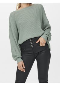 Světlý zelený oversized pletený svetr s volnými rukávy, kombinovaný s černými skinny jeans s vysokým pasem a kovovými knoflíky.