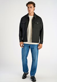 Svart skjorta med knappar och två bröstfickor över en vit skjorta, i kombination med blå denimjeans och svarta läderskor. Enkel och ren design.