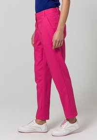 Osoba ubrana w jaskrawo różowe spodnie i białe sznurowane buty z różowymi akcentami, stojąca na tle jednolitego jasnego tła.