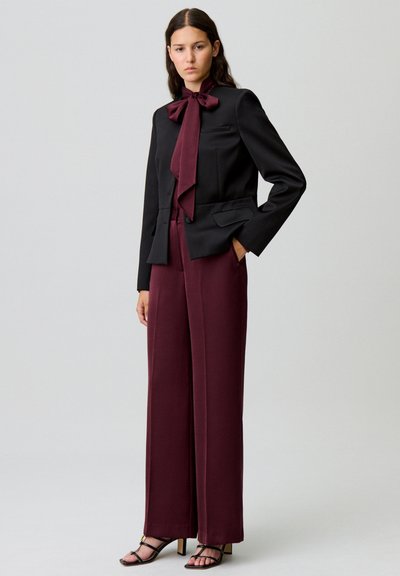 Blazer nero con dettaglio a fiocco, abbinato a pantaloni larghi bordeaux. Modella indossa tacchi neri con cinturini, in piedi contro uno sfondo neutro.
