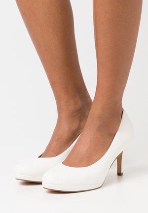 Chaussures à talons hauts blanches avec un bout rond, une surface en cuir verni lisse, et un talon fin, présentant un design échancré autour de la cheville.