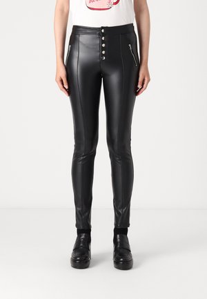 Kobieta nosząca czarne spodnie z wysokim stanem wykonane ze sztucznej skóry, z przodu zapinane na guziki, z kieszeniami na zamek błyskawiczny, czarne mokasyny oraz białą koszulkę z nadrukiem.