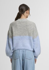 Egy sötét hajú nő, akinek alacsony lófarmer van, szürke és világoskék vastag kötött pulóvert és világoskék magas derekú farmert visel, háttal áll.