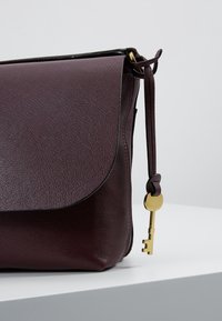 Bordeaux kožená taška přes rameno s klopnou, zdobená akcentem ve tvaru klíče ve zlaté barvě připevněným na poutku. Texturovaný povrch a elegantní design.